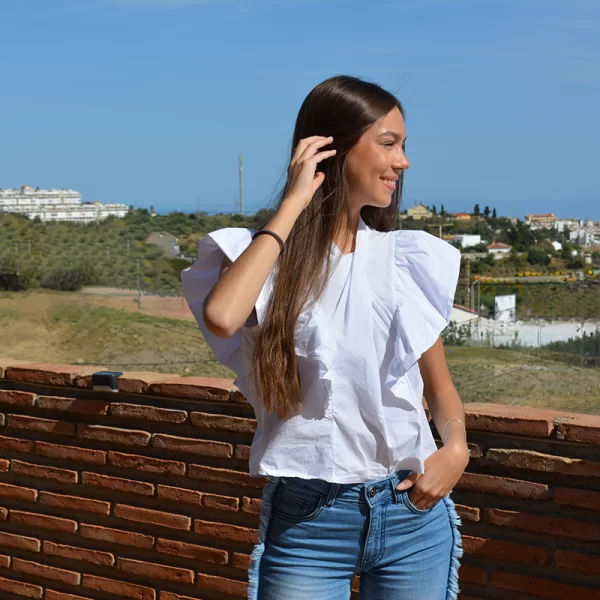 chica sonriendo con una mano en el pelo con una Blusa blanca con volantes en manga corta y tejano azul con flecos en los laterales