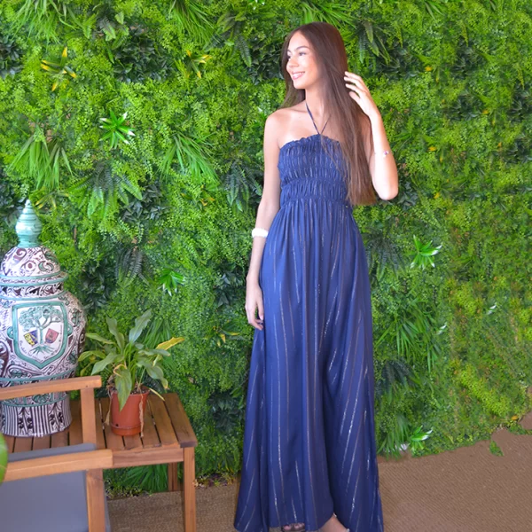 chica posando delante de una pared de flores verdes con un vestido largo palabra de honor en color azul marino con hilo de lurex y fruncido en el pecho. Cordón en el cuello.