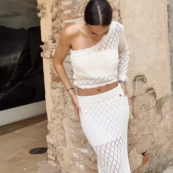 chica delante de una pared con un conjunto blanco de crochet con parte de arriba asimétrica con manga larga y falda larga con forro interior
