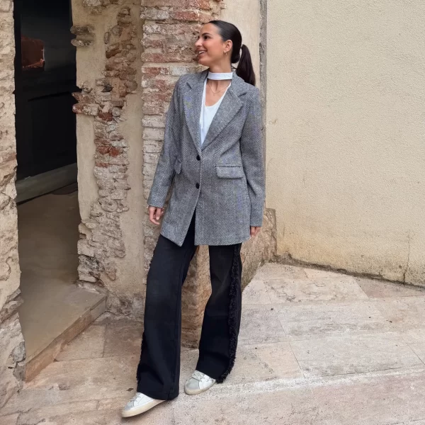 chica sonriendo en la calle con un abrigo de espiga negro con 2 botones, blusa blanca choker y tejano negro con desflecados laterales