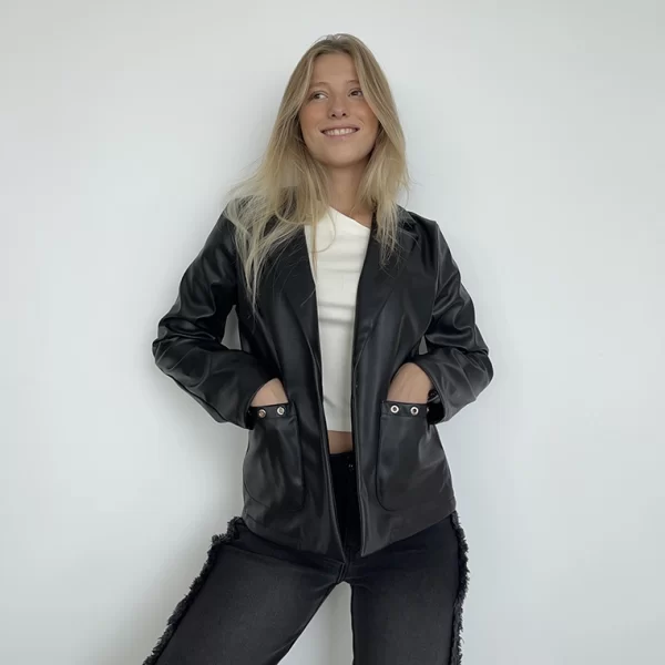 chica rubia apoyada en una pared blanca vestida con un top balnca, blazer de polipiel negro con dos bolsillos con arandelas plateadas y pantalón negro con flecos en los laterales