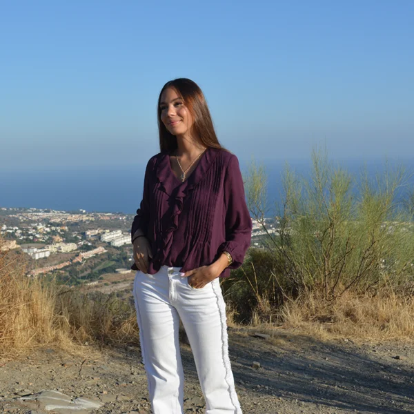 chica posndo en el campo con una blusa de bambula en color burdeos con volante en el frontal y tablitas a cada lado del volante con un tejano blanco