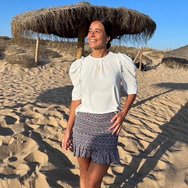chica en una playa de arena fina y dorada con una sombrilla detrás vestida con una blusa blanca de mangas francesas abullonada y falda corta fruncida con gomas elásticas estampada