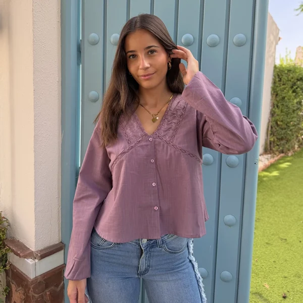 chica posando con una blusa color berenjena con escote pico y detalles bordados alrededor de escote con entredóa en cuerpo y cierre frontal con botones. Lleva un pantalón tejano azul con flecos en los lados