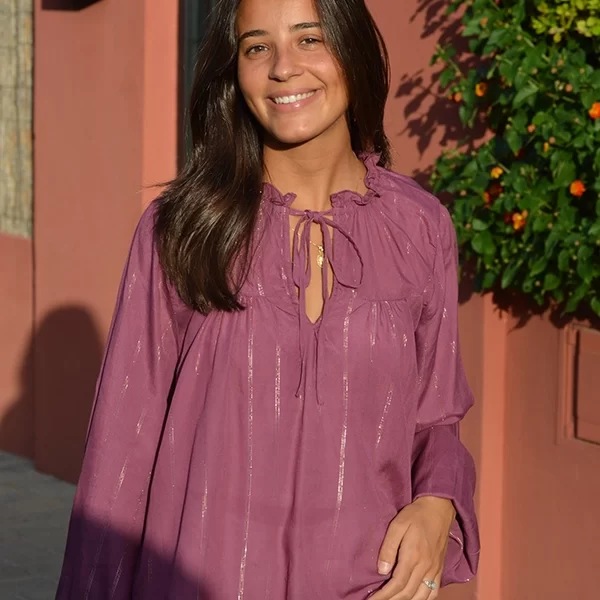 chica sonriendo delante de un muro de una casa con una blusa amplia rosa viejo con líneas de lúrex doradas y plateadas