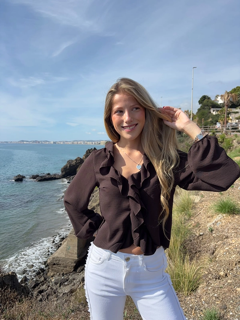 chica rubia posando en un acantilado con una blusa de color marrón chocolate con un volante alrededor del cuello y frontal
