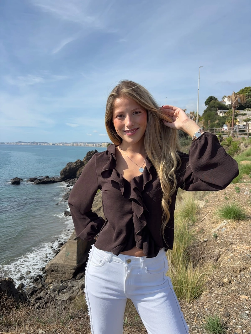 chica rubia posando en un acantilado con una blusa de color marrón chocolate con un volante alrededor del cuello y frontal