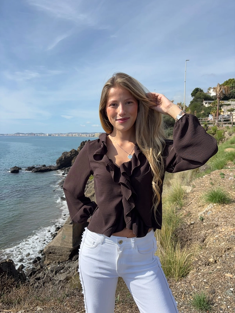 chica rubia posando en un acantilado con una blusa de color marrón chocolate con un volante alrededor del cuello y frontal