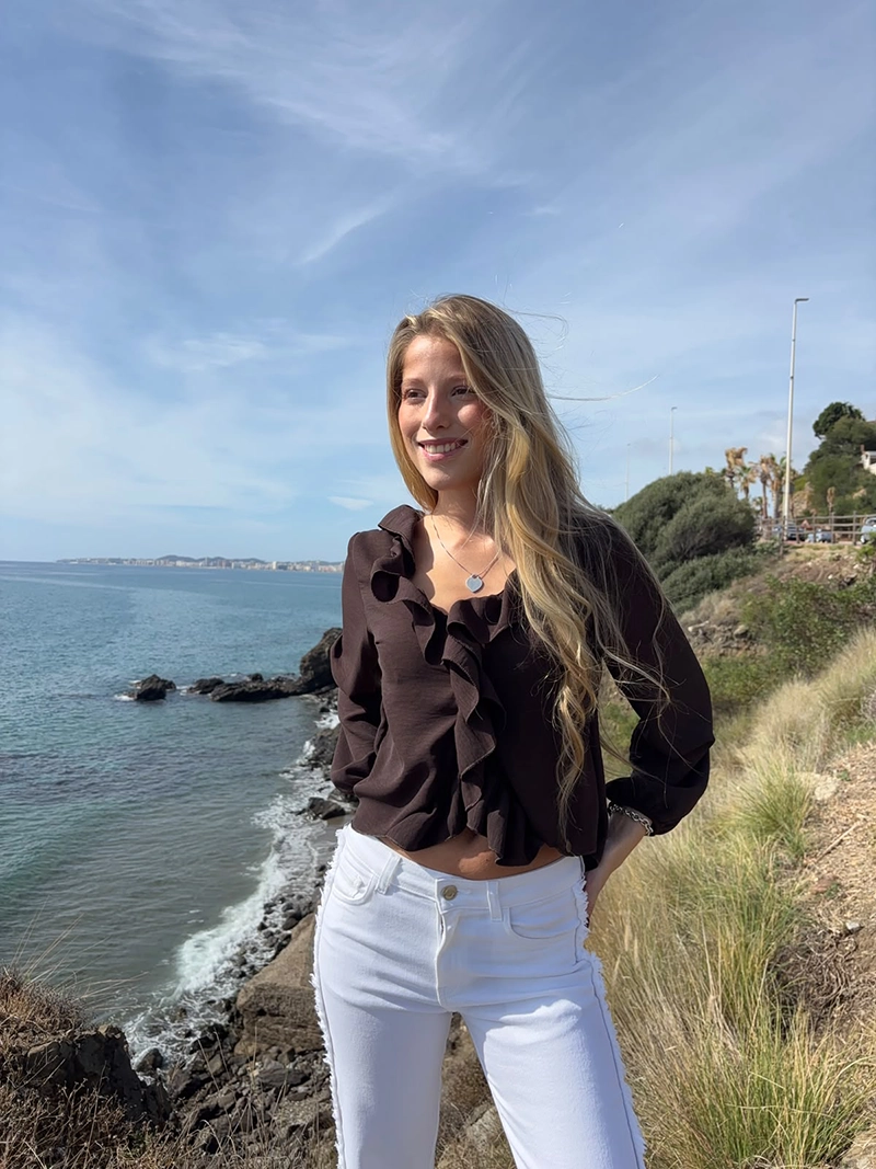 chica rubia posando en un acantilado con una blusa de color marrón chocolate con un volante alrededor del cuello y frontal