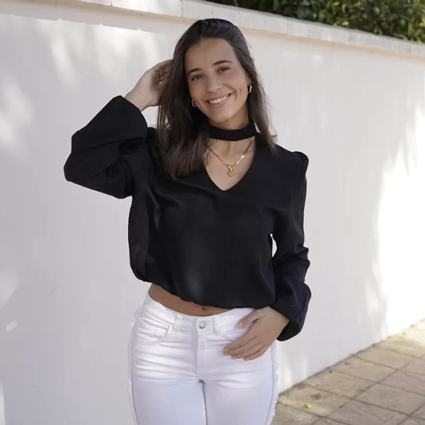chica sonriendo con una blusa negra de manga larga, cuello pico y choker y pantalón blanco