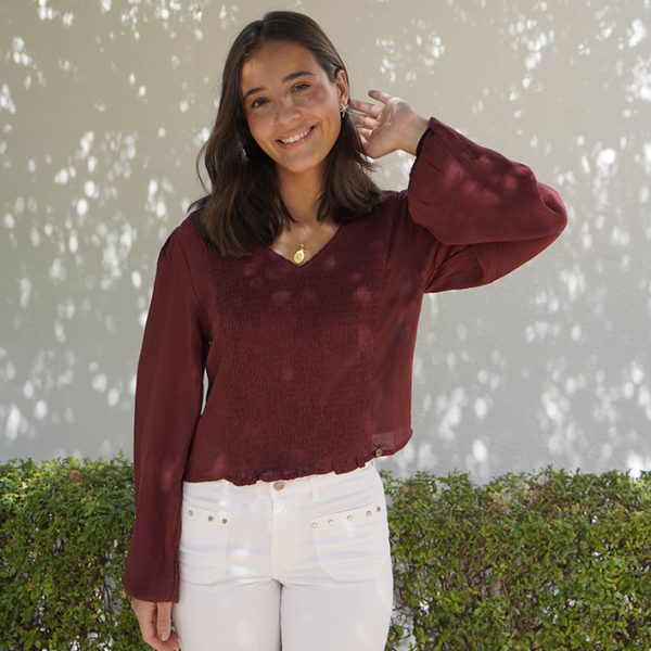 chica posando de pie, sonriendo con una mano en el pelo vestida con una blusa burdeos con fruncido en la parte central del frontal, escote pico y manga larga