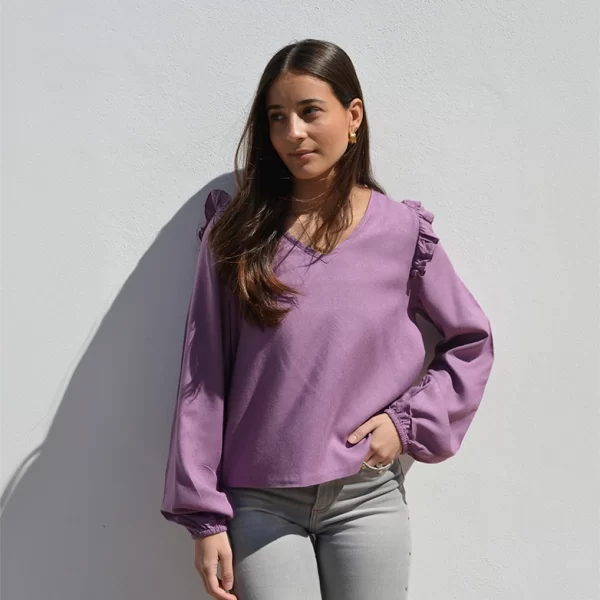 chica apoyada en una pared lisa gris con una blusa de manga larga rosa con colantes en hombros y pantalón gris con tachuelas plateadas en bolsillos y laterales