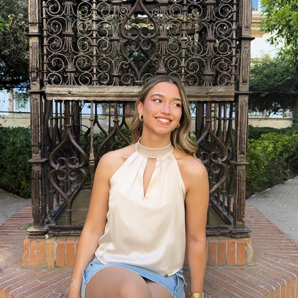 chica sentada en un parque sonriendo vestida con una blusa dorada con cuello halter y falda cargo tejana azul