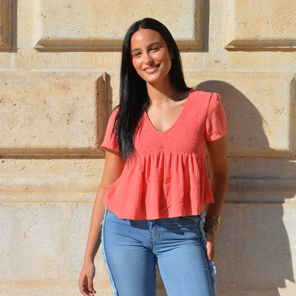 chica posando con una blusa manga corta fruncida en la parte de arriba en coral con un pantalón tejano azul claro desflecado en los laterales