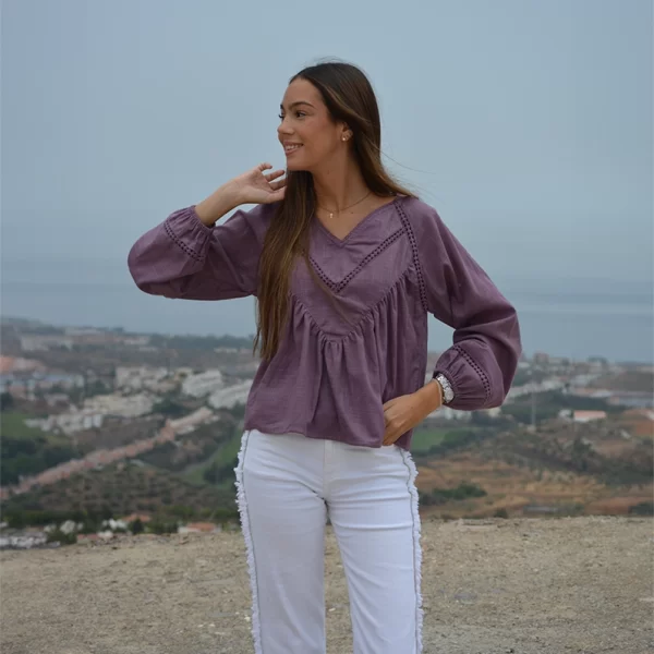 chica sonriendo mirando a la derecha con una mano en la cintura con una blusa berenjena con tira bordada decorativa. Forro en el cuerpo y goma elástica en puños. 100% Algodón y un pantalón denim blanco con flecos a los lados
