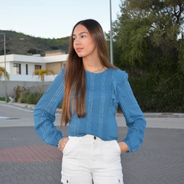 chica en una calle con una blusa azul de manga larga, cuello redondo, volantes en hombros y rayas de lurex en plateado con pantalón blanco cargo
