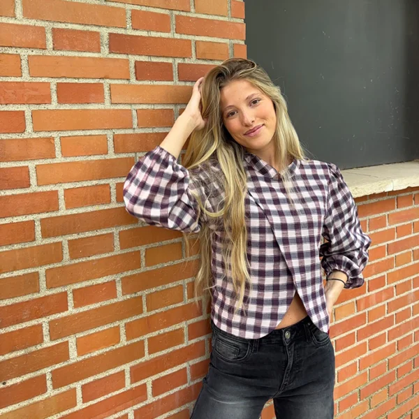 chica posando en una pared de ladrillos con una blusa abierta cruzada frontal de cuadros burdeos y pantalón negro