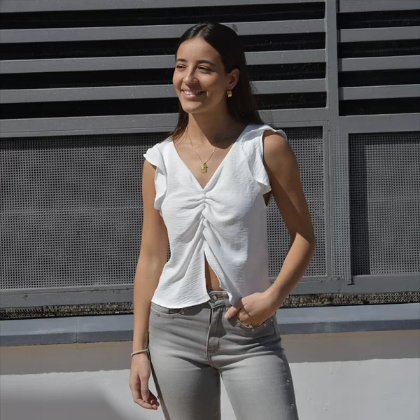 chica en la calle con un pantalón gris claro con tachuelas en bolsillos y laterales y blusa blanca sin manga, con un pequeño volante en hombro y abertura en bajo frontal central. escote en v