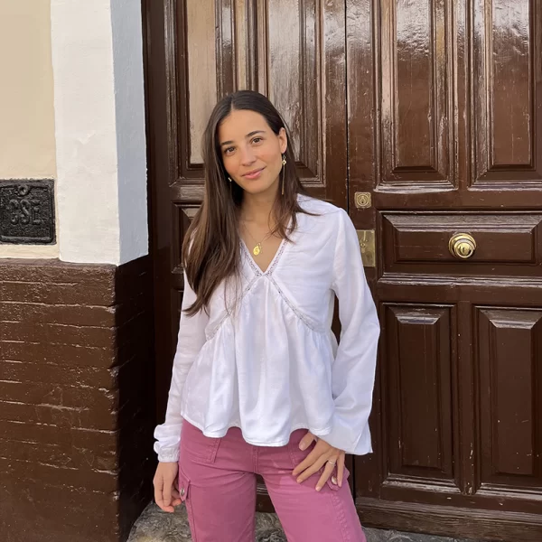 Chica joven posando delante de una puerta de madera marrón con una blusa blanca con escote pico y amplia en el bajo, con intersecciones en costuras frontales y cuello y un pantalón cargo rosa