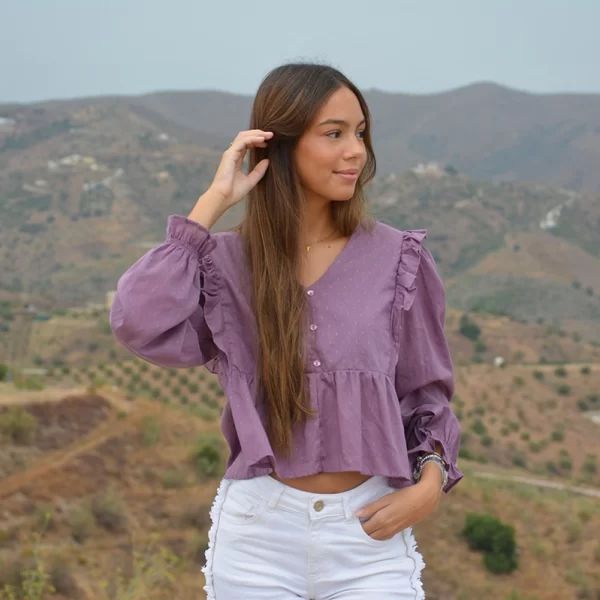 chica sonriendo con una blusa plumeti en color berenjena con escote pico, botones decorativos, volantes en frontal y trasero de ambos laterales y goma fruncida en puños, con un pantalón denim blanco con flecos en laterales