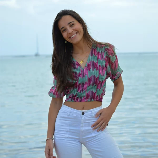 chica posando para un foro delante del mar con una blusa cruzada de gasa con estampado en tonos verdes, malvas y rosas con hilo de lúrex plateado. Manga francesa abullonada y un pantalón blanco