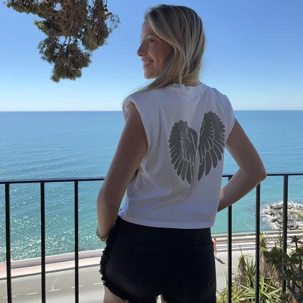 chica posando delante del mar con una camiseta blanca con un estampado de alas en color plateado en la espalda