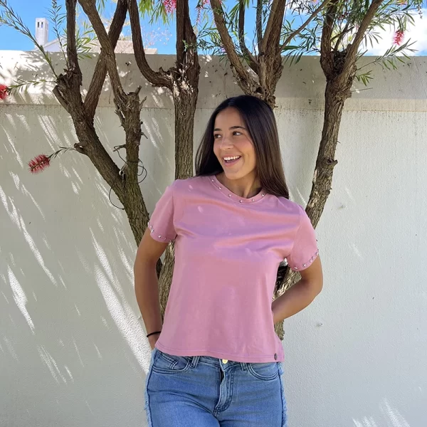chica posando en jardin con una camiseta de manga corta en color rosa con tachuelas plateadas bordeando el filo del cuello y las mangas
