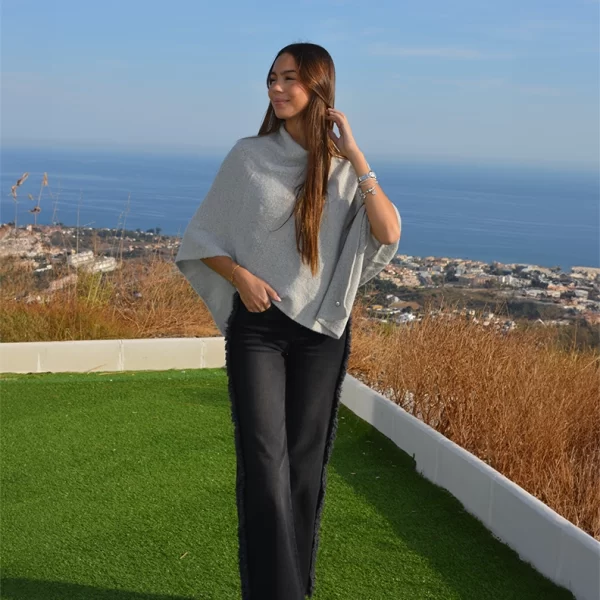 chica posando en lo alto de un monte con vistas al mar y montaña con una mano en el pelo vestida con una capa gris jaspeada y pantalón negro con flecos en laterales