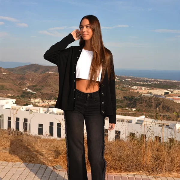 chica posando en lo alto de un monte con vistas al mar y montaña con un cárdigan negro con arandelas plateadas alrededor de la abertura frontal, un top corto blanco y un pantalón negro con flecos en los lados