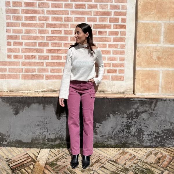 chica posando en la calle delante de una pared de ladrillos rojizos pequeños con un jersey blanco con hombro izquierdo descubierto y cuello choker. Los puños son ligeramente acampanados y lo lleva con un pantalón cargo rosa