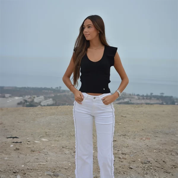 chica posando con un Top de tricotosa en color negro con mangas de volantes y pantalón denim blanco con flecos a los lados