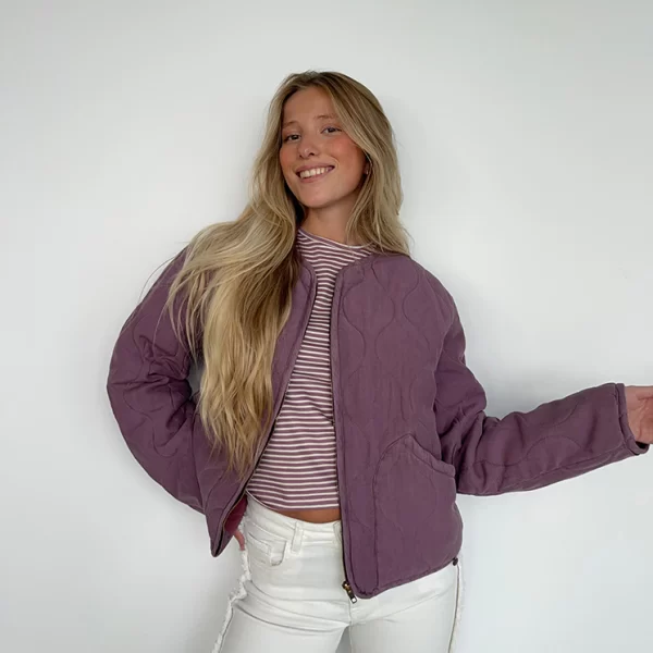 chica posando con una camisetas de rayas berenjena, pantalón blanco con flecos laterales y chaqueta acolchada rosa viejo con cremallera frontal