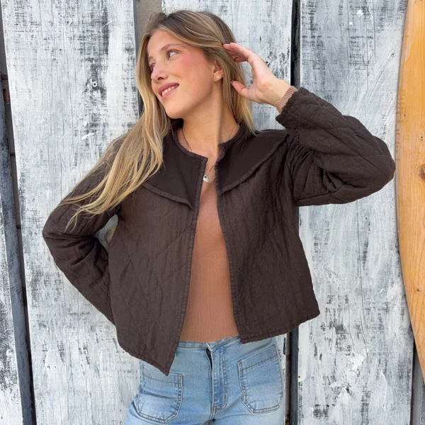 chica rubia posando delante de unos tablones grises con una chaqueta fina de color marrón