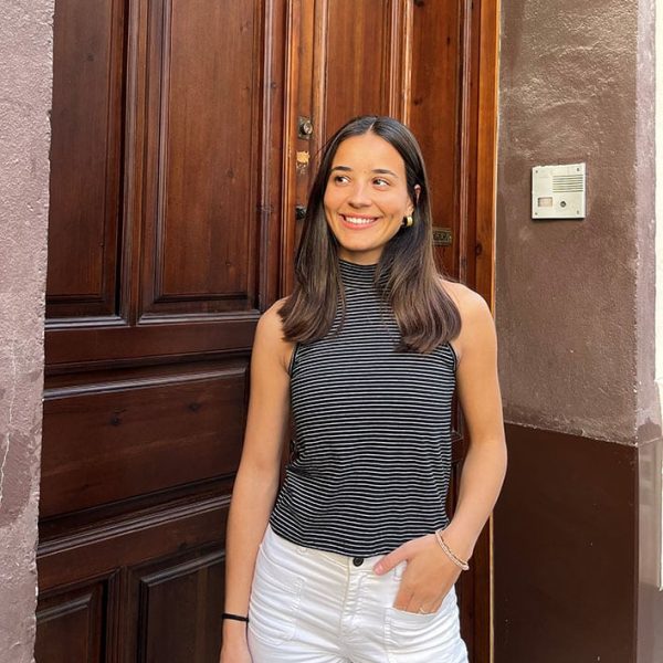 chica sonriendo delante de una puerta de madera con una mano en el bolsillo del pantalón blanco y con un top sin mangas negro de rayas finas blancas y sin mangas