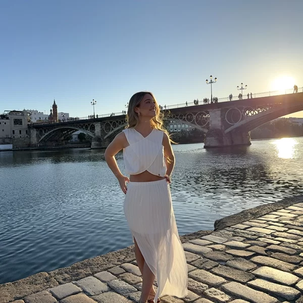 chica posando en Sevilla, delante del río Guadalquivir con un Conjunto de blusa y falda blanca con hilos de lúrex plateados y dorados. Blusa cruzada con forro interior en la parte frontal cruzada Falda larga con aberturas en ambos bajos laterales y forro corto interior