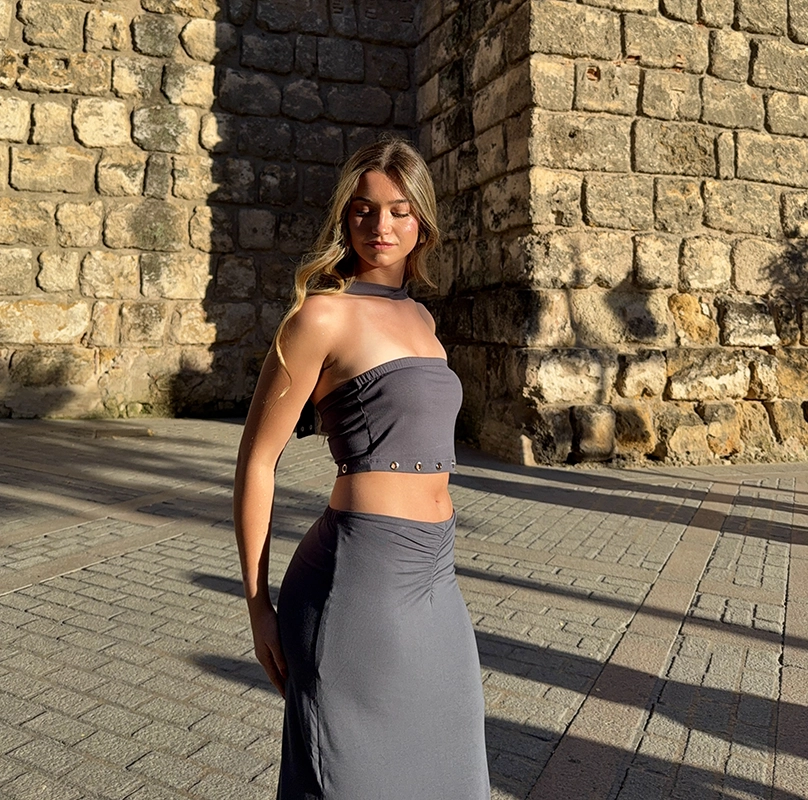 chica posando con Set de viscosa elástica en color antracita con aros metálicos en color plata en el top y en los extremos de la cinta del cuello. Falda con frunce en en frontal de la cintura y aberturas laterales.