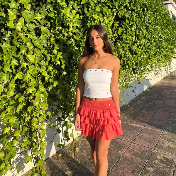 chica paseando con una Falda pantalón coral anaranjado con cuatro volantes, fruncido con gomas elásticas en cintura y short interior de punto liso a tono y top palabra de honor blanco con tachuelas en escote