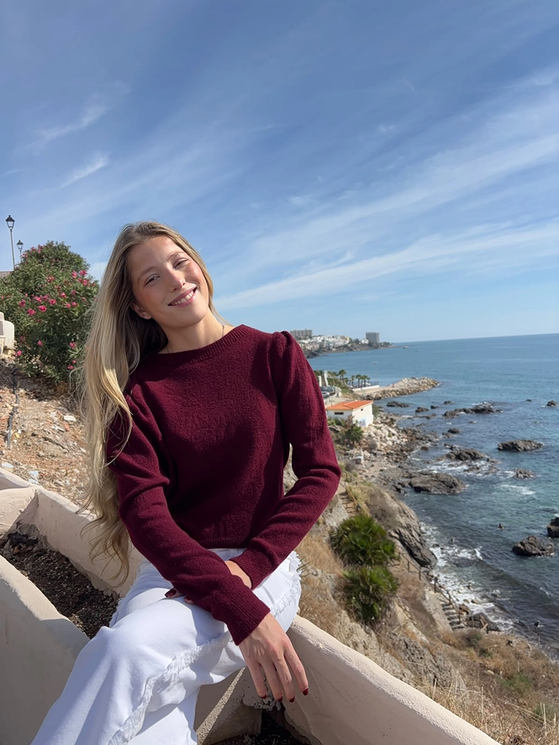 chica rubia sentada posando en un acantilado frente al mar con un jersey burdeos con los hombros fruncidos