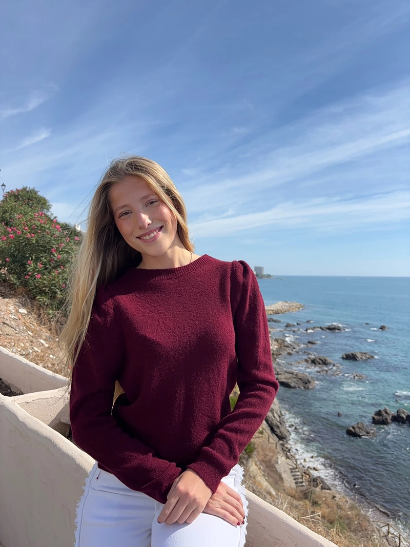 chica rubia sentada posando en un acantilado frente al mar con un jersey burdeos con los hombros fruncidos