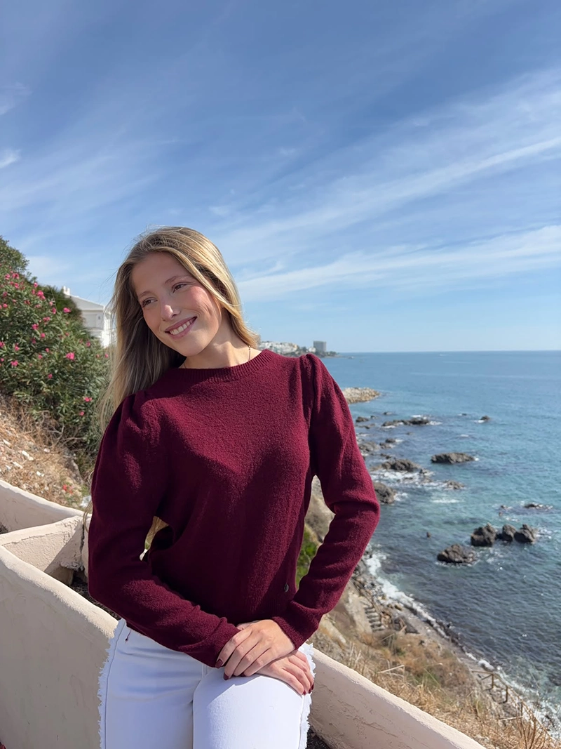 chica rubia sentada posando en un acantilado frente al mar con un jersey burdeos con los hombros fruncidos
