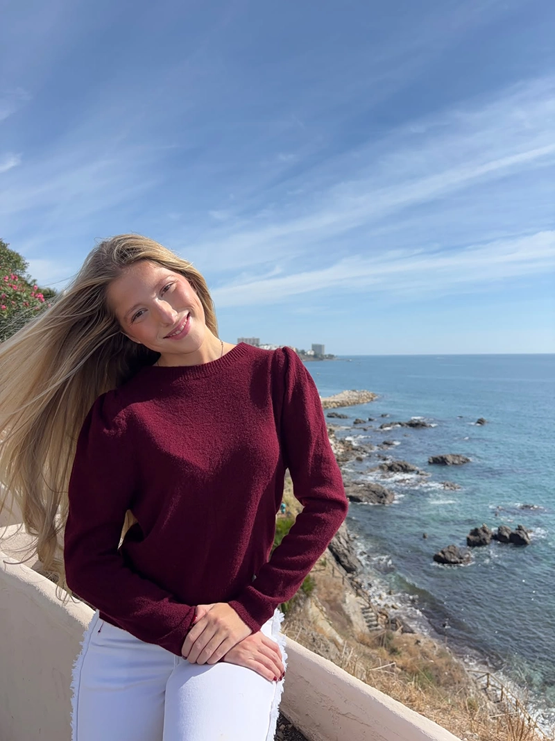 chica rubia sentada posando en un acantilado frente al mar con un jersey burdeos con los hombros fruncidos