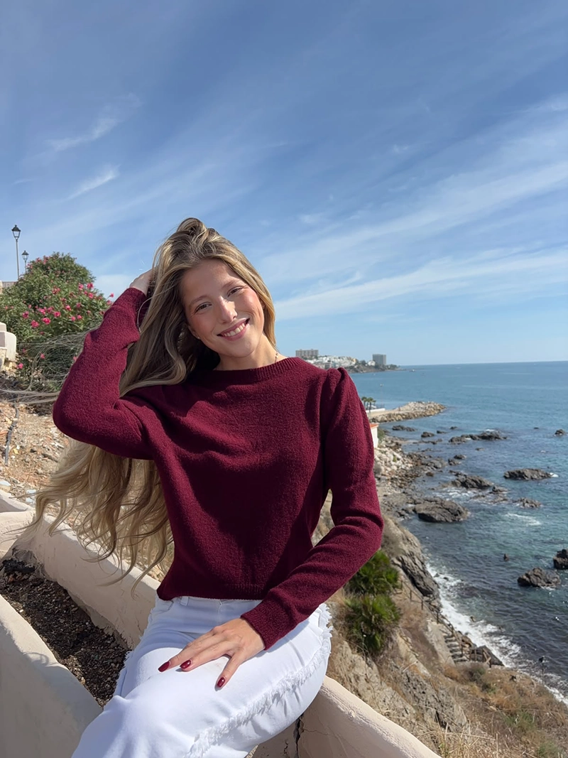 chica rubia sentada posando en un acantilado frente al mar con un jersey burdeos con los hombros fruncidos