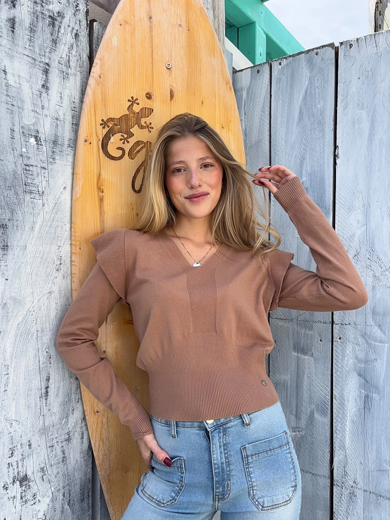 chica rubia posando delante de una tabla de surf de madera con un jersey camel con volantes en los hombros