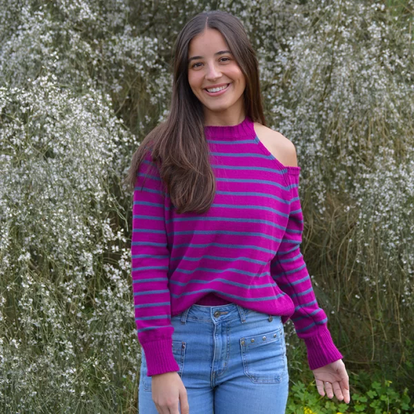 chica sonriendo de frente en el campo andando con un jersey tricotosa de color fucsia orquídea con rayas en color azul y con hombro izquierdo descubierto. Detalle de chapa metálica Pink Pomelo en el bajo lateral izquierdo.