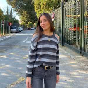 Chica con pelo castaño suelto que viste un jersey de rayas con tres colores de grises con aberturas en ambos hombros.