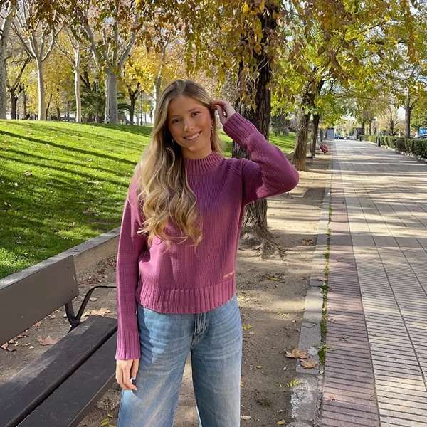 chica rubia en un parque con un pantalón tejano azul claro y un jersey rosa con un bordado en color maiz en el bajo frontal izquierdo que pone pink pomelo