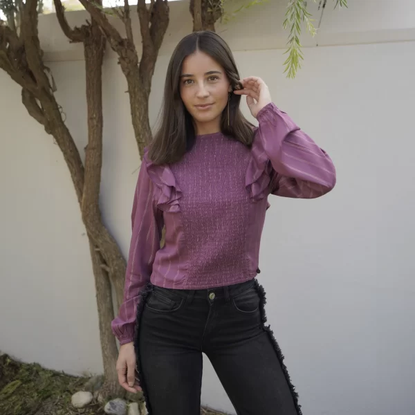 chica posando con una blusa fruncida en color berenjena con líneas de lúrex doradas. Cierre en cuello trasero con botón; volantes en hombros y goma elástica en puños y pantalón negro con flecos en los laterales
