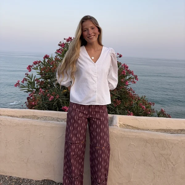 chica sonriendo delante del mar con un pantalón estampado en tonos burdeos y una blusa blanca plumeti con manga 3/4 en color blanco
