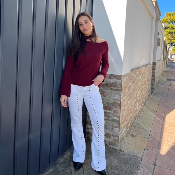 chica delante de una puerta antracita con un pantalón blanco con dos líneas frontales de tachuelas y un jersey asimétrico con cuello choker en burdeos