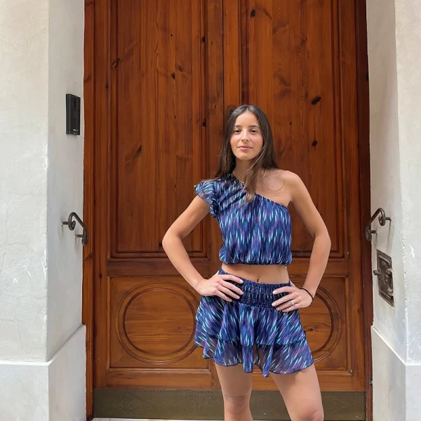 chica delante de una puerta marrón con una blusa asimétrica estampada en tonos azules y malvas con goma elástica suave en cintura escote y 2 volantes en hombro derecho y falda pantalón del mismo tejido de la blusa